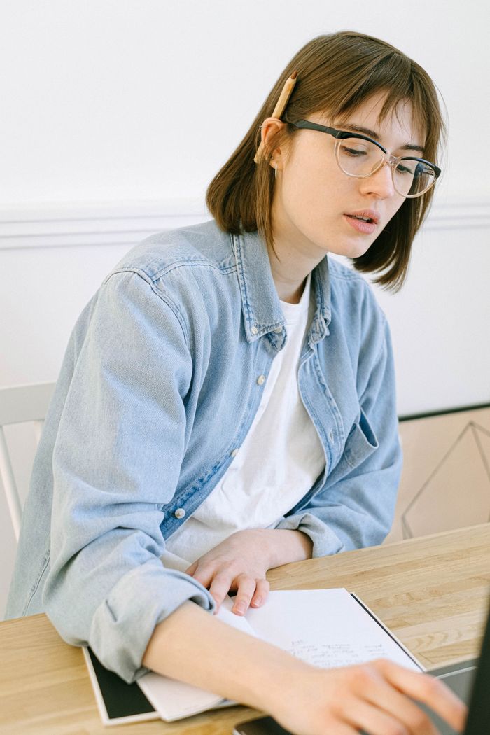 Offerings Young woman in casual attire writing notes at a home office desk, concentrating intently.